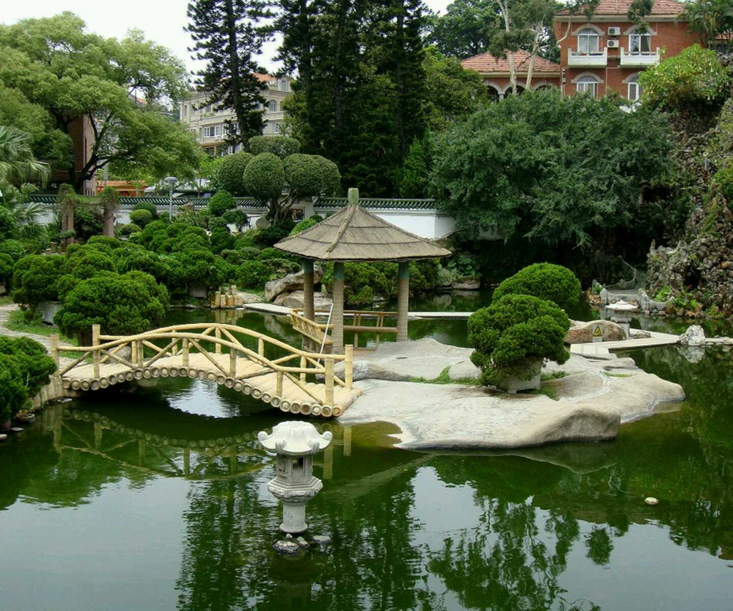 Tranquil garden pond with a bamboo bridge leading to a thatched-roof pavilion, surrounded by manicured shrubs