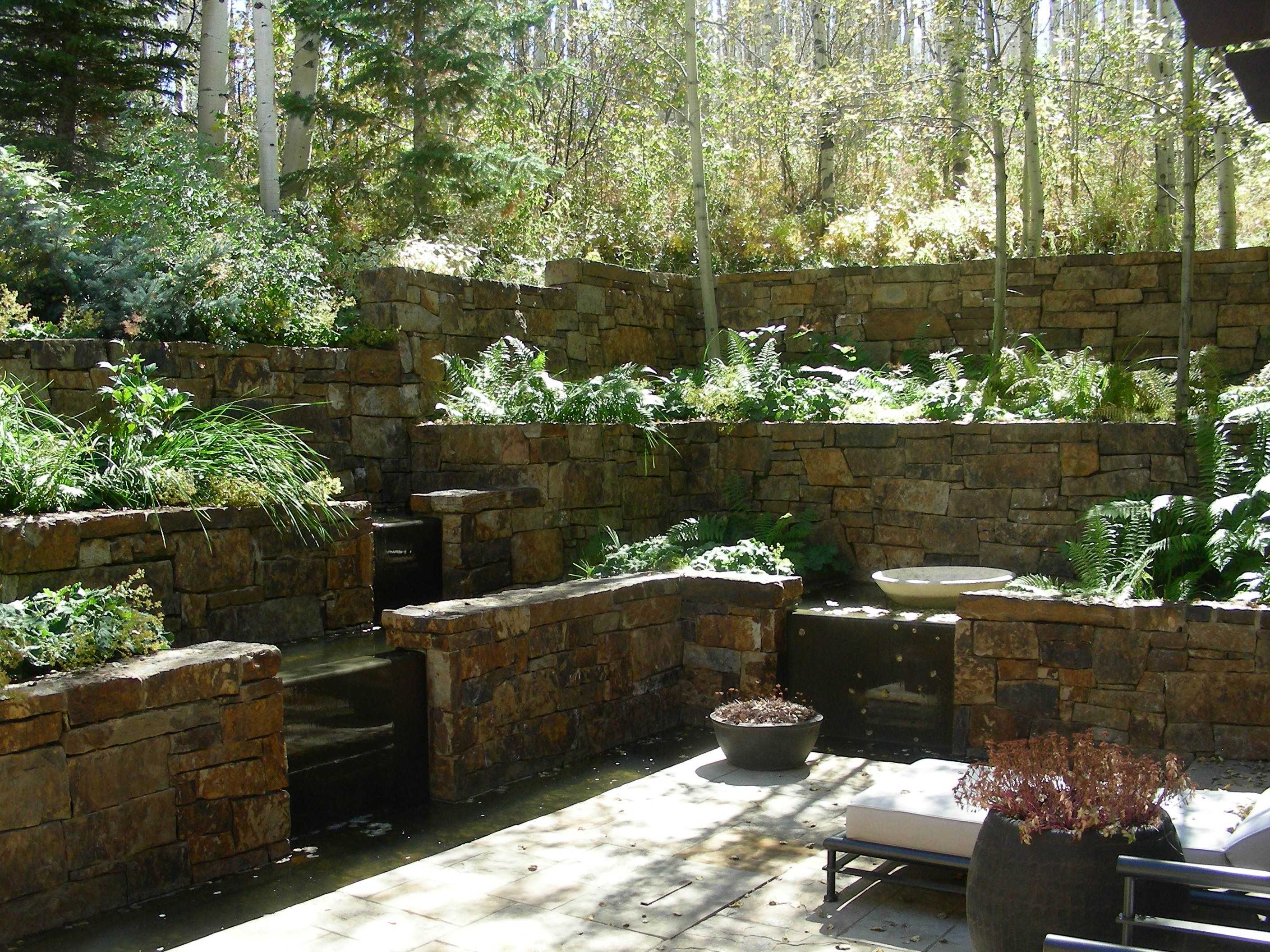 Tiered stone retaining walls with ferns and greenery, a small water feature, and outdoor seating in a wooded garden setting