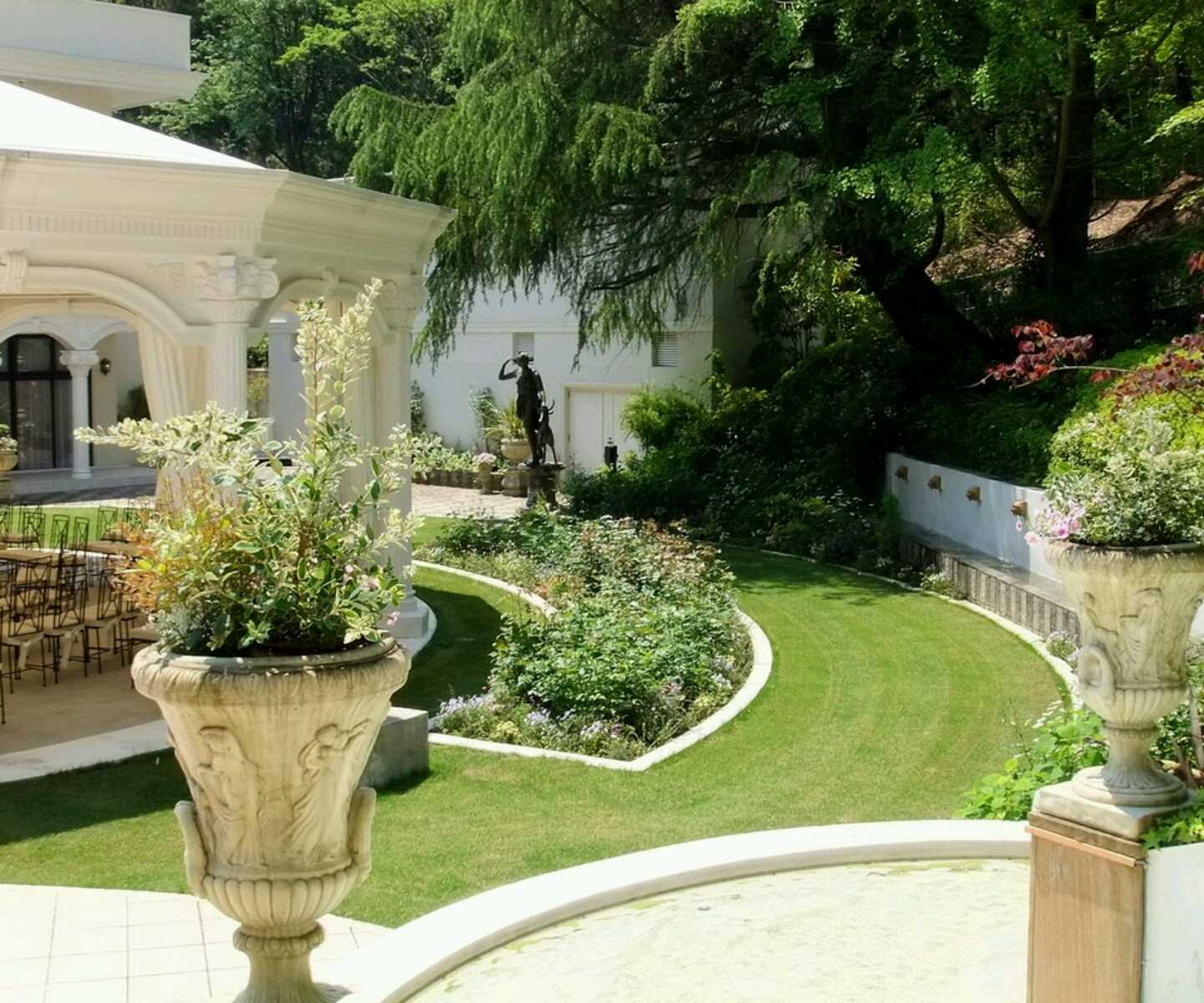 Elegant garden courtyard with ornate stone urns holding plants, manicured lawn, flower beds, a bronze statue, and a white building